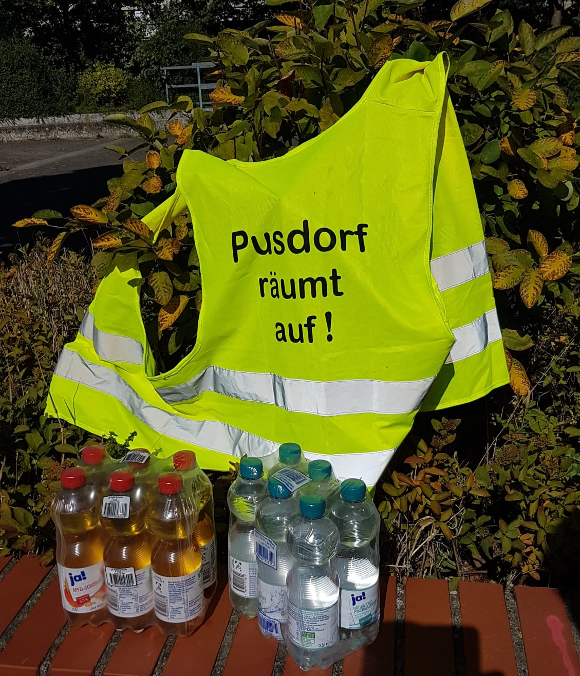 eine Warnweste von Pusdorf räumt auf hängt in einer Hecke. Auf einer kleinen Steinmauer darunter stehen Plastikflaschen mit Wasser und Apfelschorle