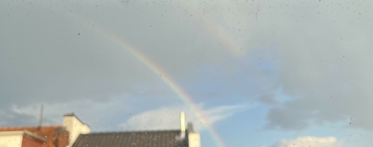 a blurry shot of a rainbow above city roofs, in front of a partially blue, partially grey sky