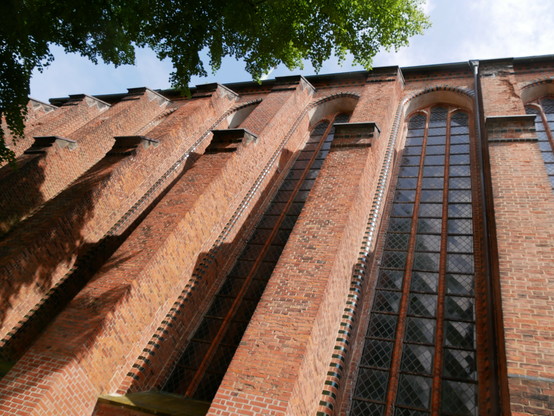 Blick nach oben auf das Seitenschiff der St. Michaeliskirche Lüneburg von außen. Vor der Kirche steht eine Linde und ein wenige leicht bewölkter Himmel ist auch zu sehen.  