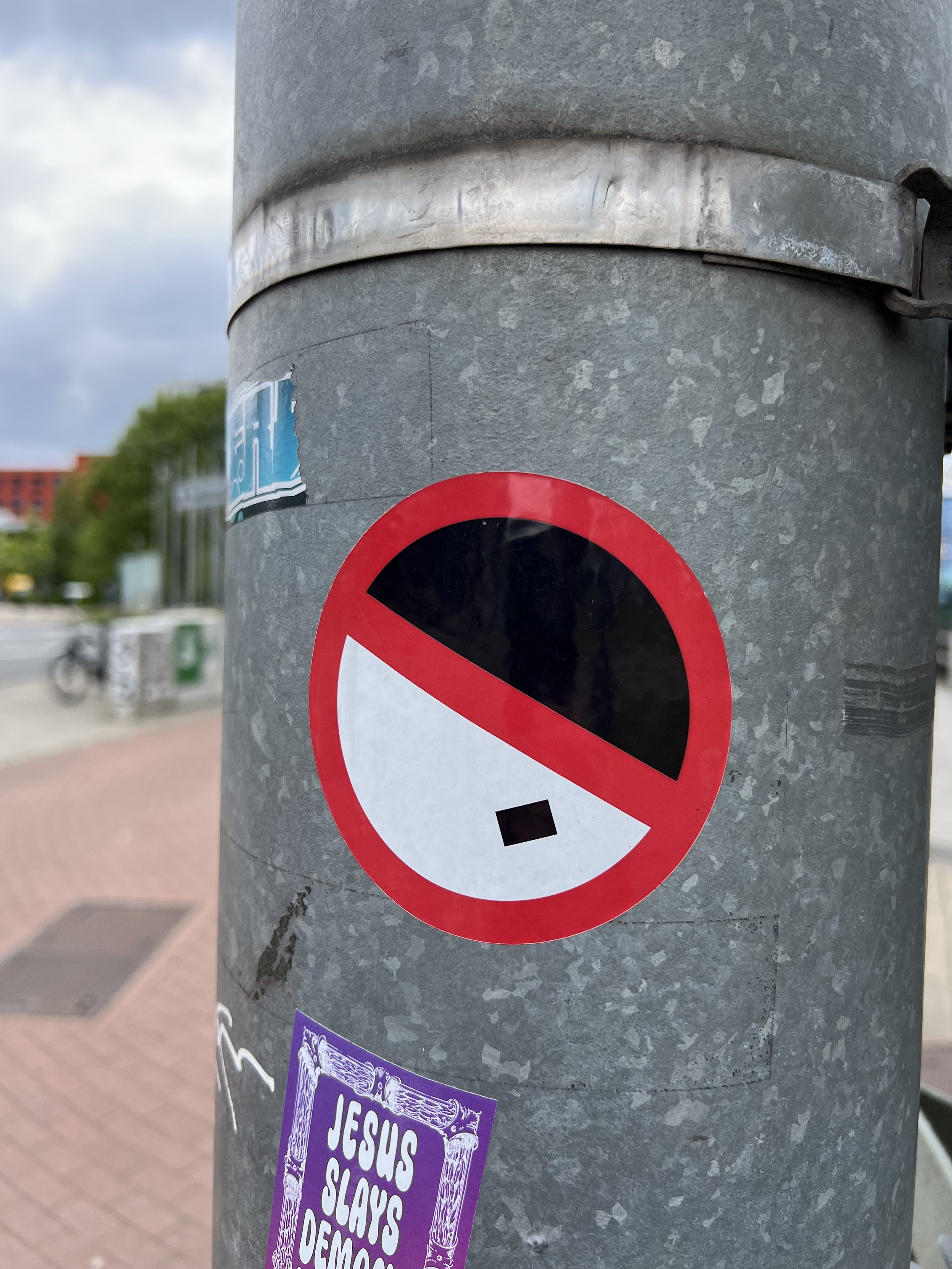 Runder Sticker an einem Laternenmast. Der Sticker hat eine rote Kontur und einen schrägen roten Balken von links oben nach rechts unten. Die obere rechte Hälfte der Form ist schwarz gefüllt, die untere linke ist weiß mit einem kleinen schwarzen Rechteck. Das ganze sieht aus wie ein durchgestrichenes Hitler-Portrait. 