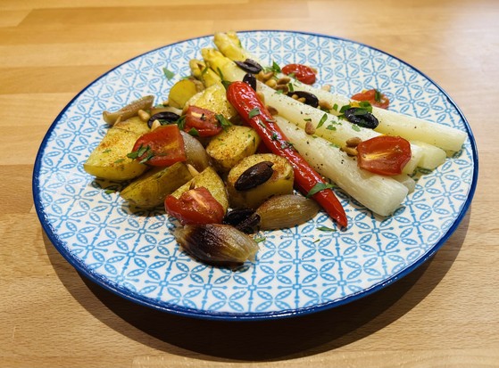 Ein Teller mit gemischtem gebratenem Gemüse, einschließlich Kartoffeln, Kirschtomaten, weißem Spargel, Paprika, schwarzen Oliven, Pinienkernen und Kräutern garniert.