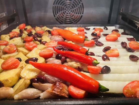 Ein Blech mit Gemüse, das im Ofen brät, darunter Kirschtomaten, rote Paprika, schwarze Oliven, Kartoffeln, Schalotten und weißer Spargel.